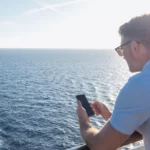 a man with a cell phone on the deck of a cruise ship looking out to the ocean.