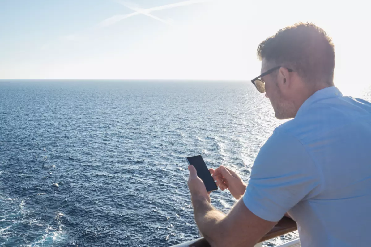 a man with a cell phone on the deck of a cruise ship looking out to the ocean.